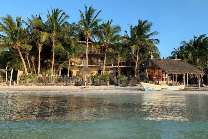 Casa Cat Ba vista desde el agua: la casa entre palmeras frente a la playa de Holbox, con una lancha en la orilla.